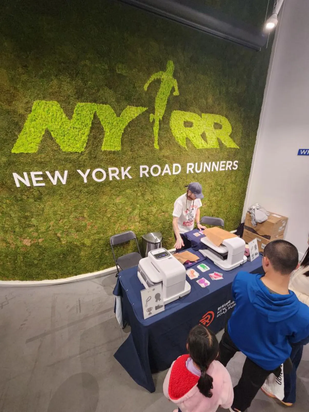 The heat press station — two presses on a draped table with design cards laid out and guests waiting — set up in front of the New York Road Runners moss wall
