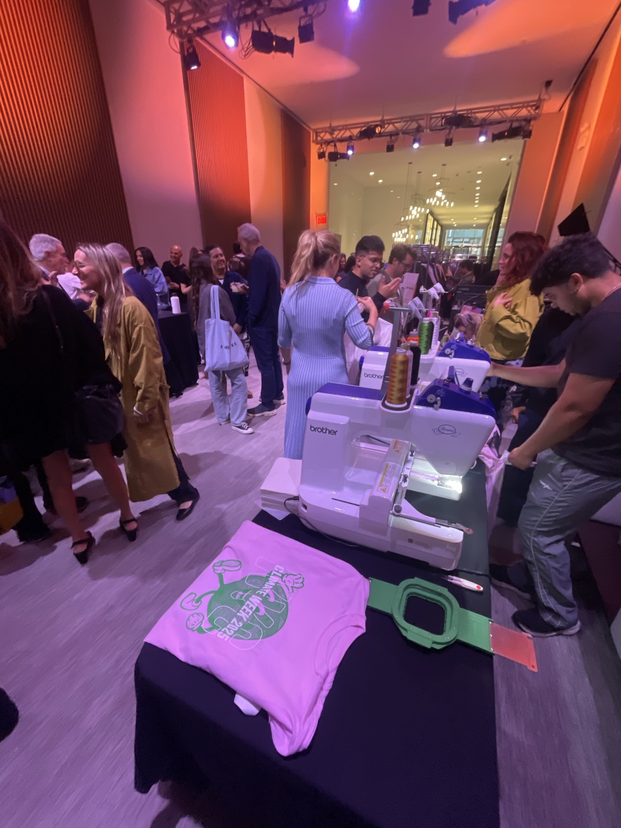 The live embroidery station mid-event at night — a finished Climate Week 2025 tee on the table, machines running and a packed room around the table
