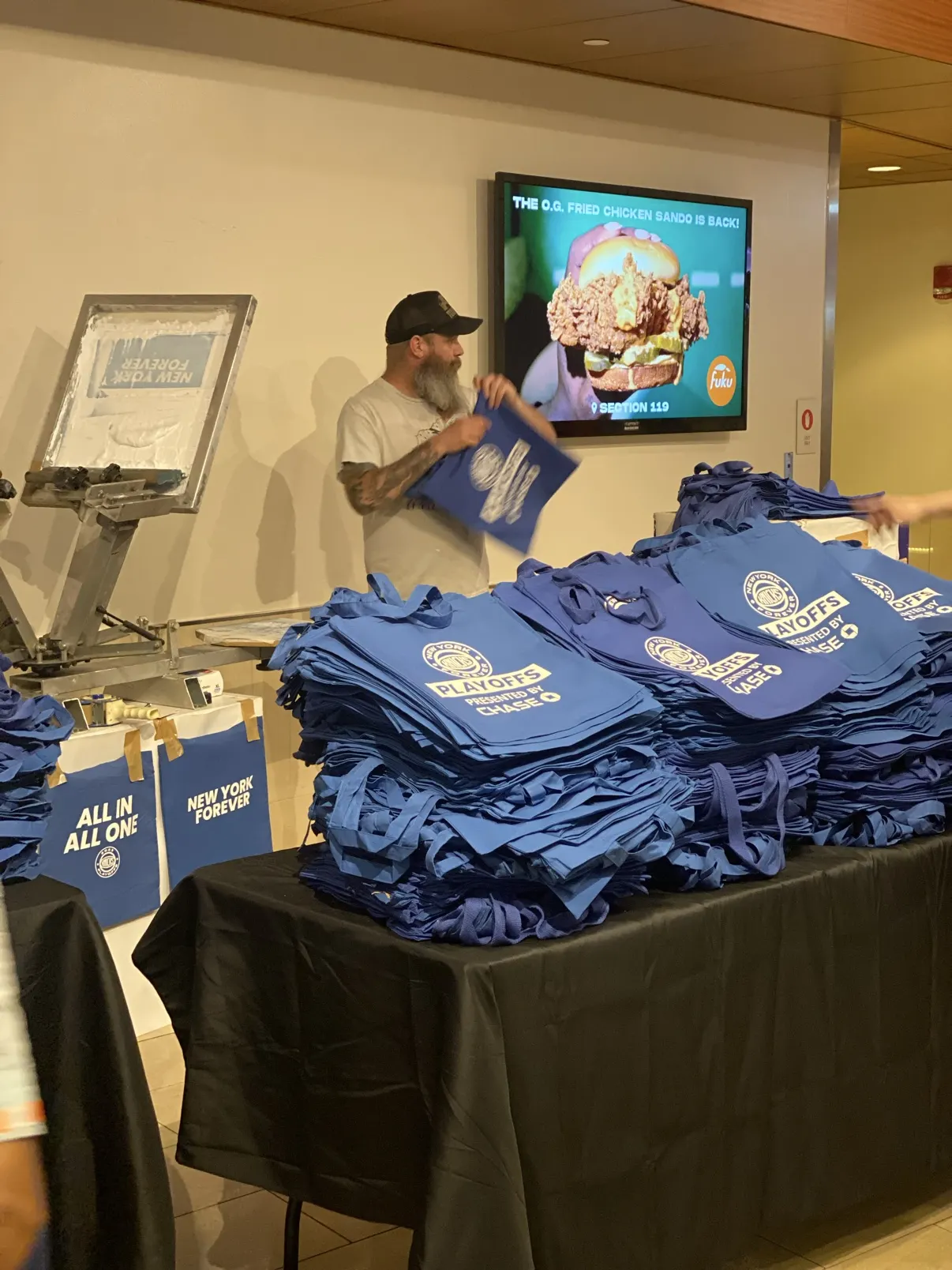 Stacks of blue Knicks Playoffs totes on the table at the arena, a crew member holding up a fresh one beside the press