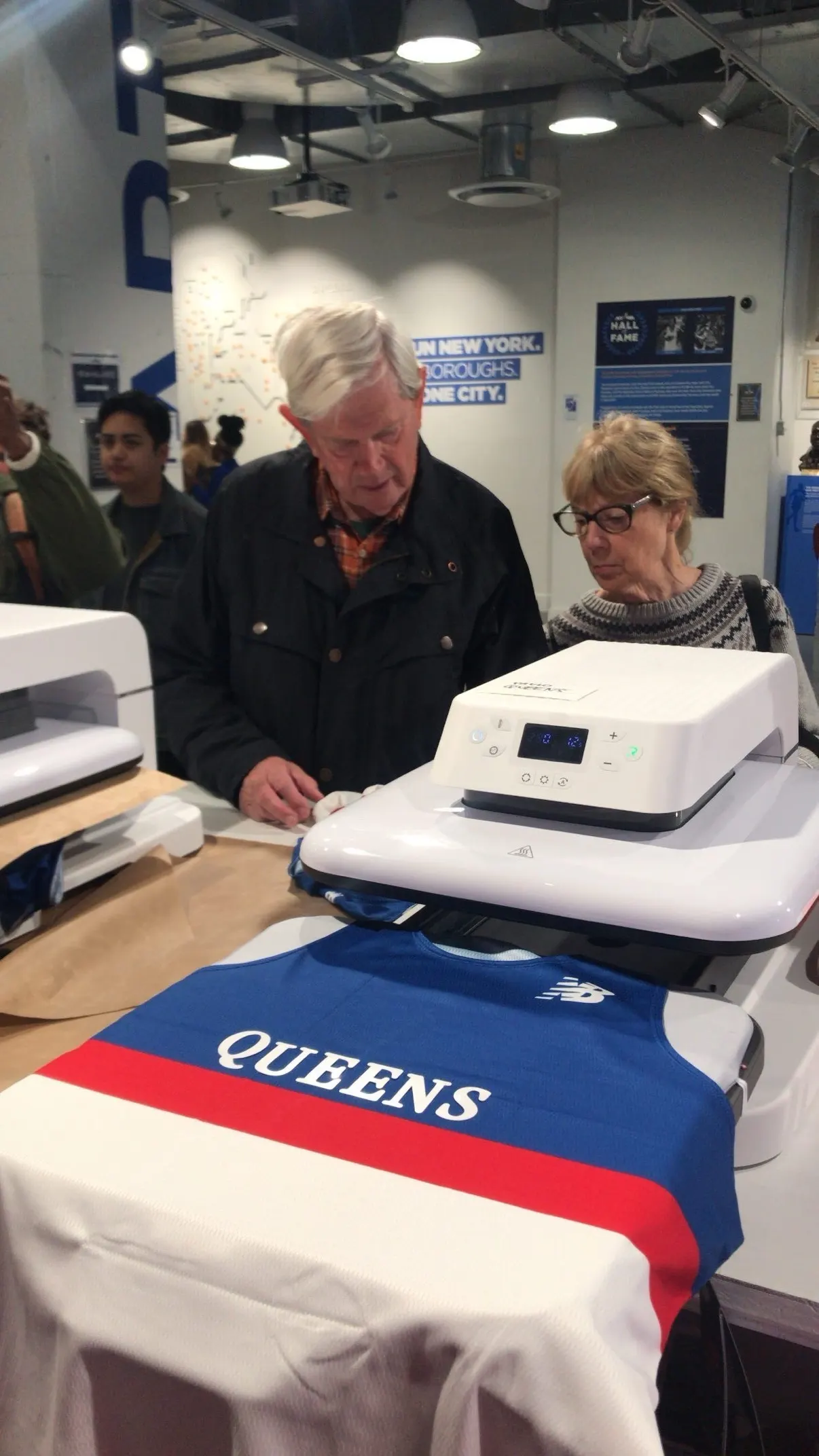 A guest's New Balance singlet reading QUEENS going under the heat press at the NYRR Run Center, a couple watching it happen