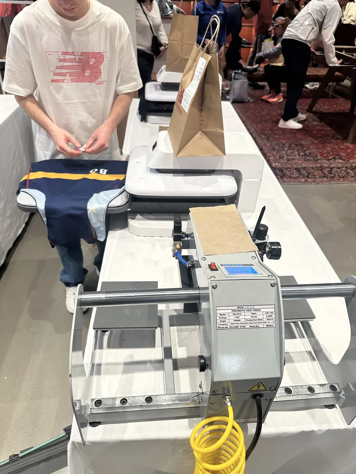 The press line on the table inside the New Balance store — pneumatic press in front, auto presses behind, a guest's singlet on the platen