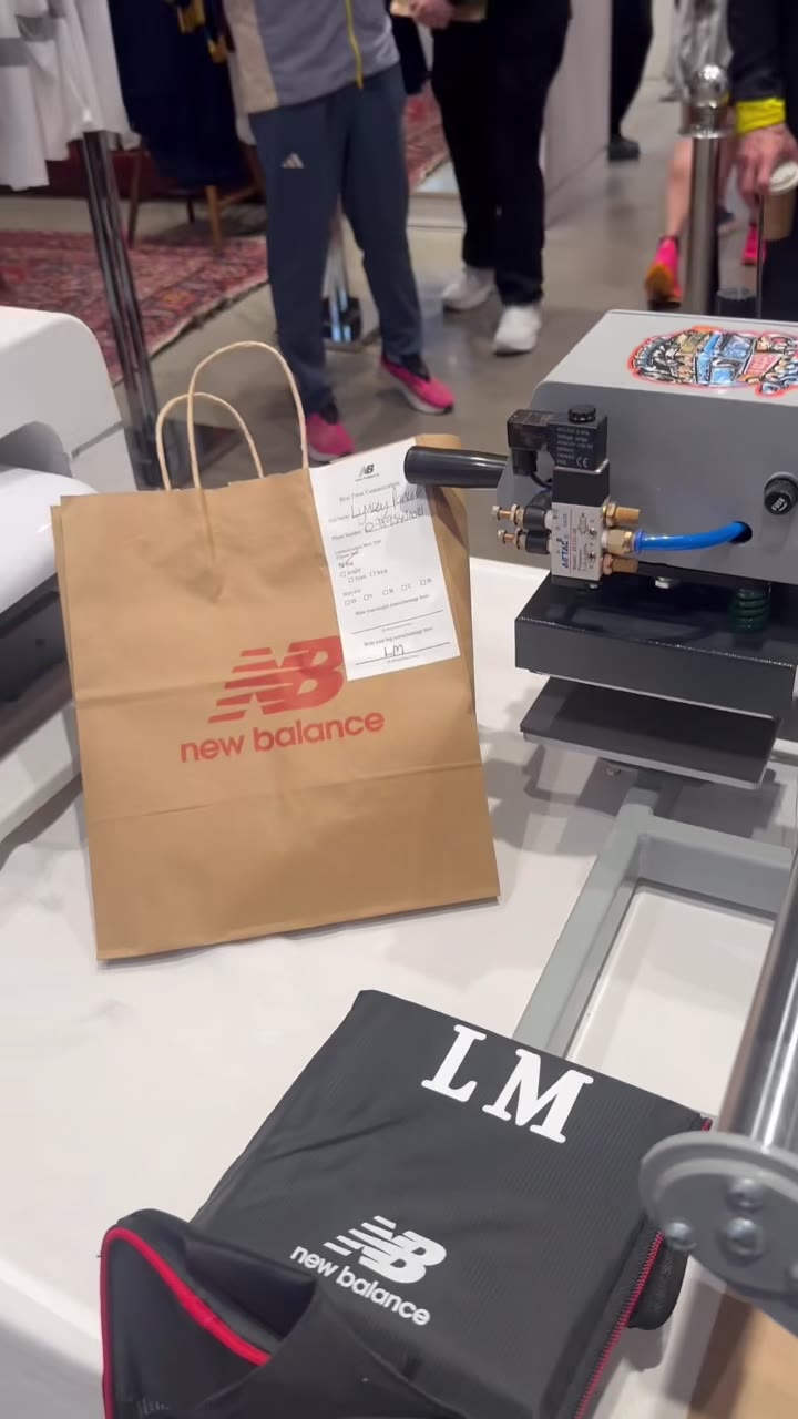 The finished New Balance shoe bag with the initials LM pressed on it, sitting next to the press beside a branded shopping bag