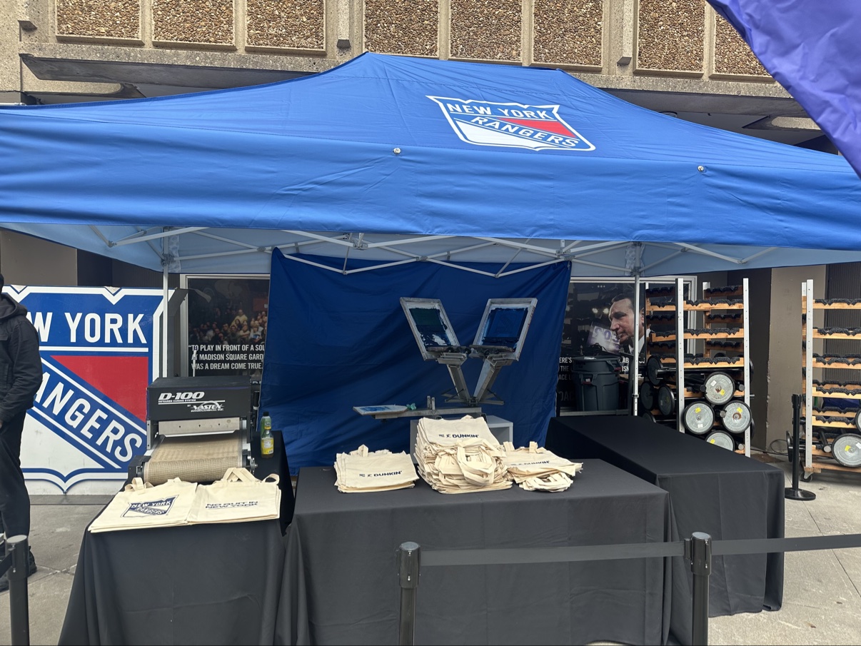 The live printing station under the Rangers-branded tent outside Madison Square Garden — press, D-100 dryer and stacks of Dunkin' × Rangers totes on the tables