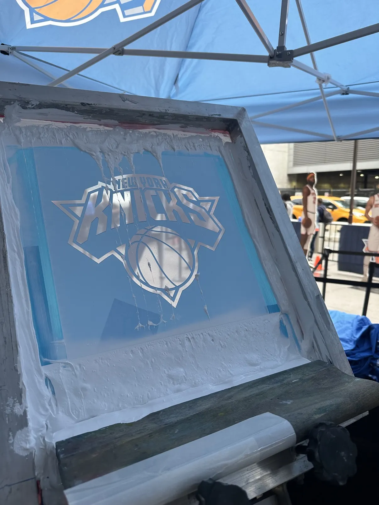 Close-up of the Knicks logo screen inked up under the tent, player standees and a yellow cab on the street behind