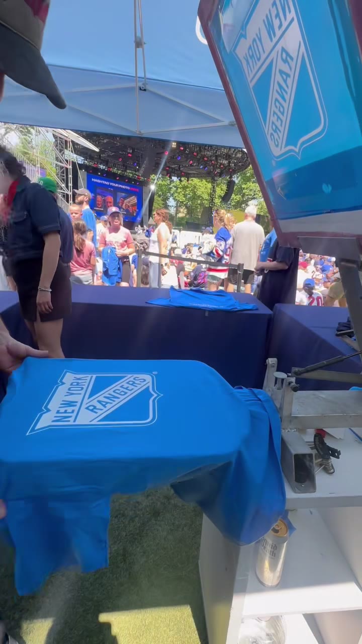 A blue Rangers shield shirt on the platen mid-print under the tent, fans gathered behind the barriers