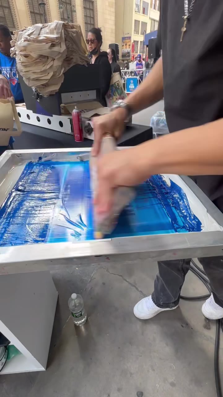 A squeegee pull in progress with blue ink at the outdoor station, a stack of totes piled behind the press
