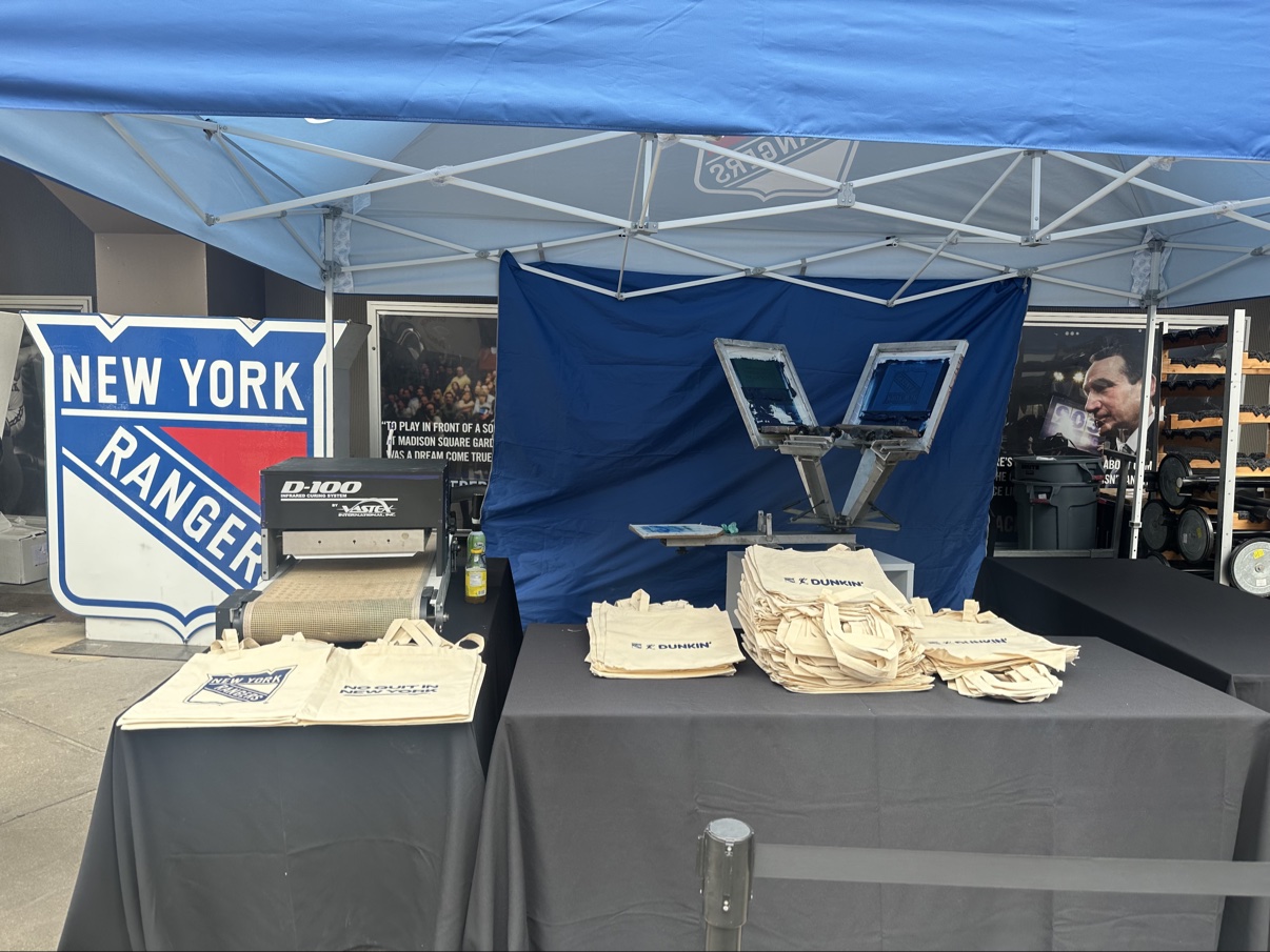 Closer view of the Rangers station outside MSG — the big shield sign, the D-100 dryer, the press and stacks of Dunkin' × Rangers totes