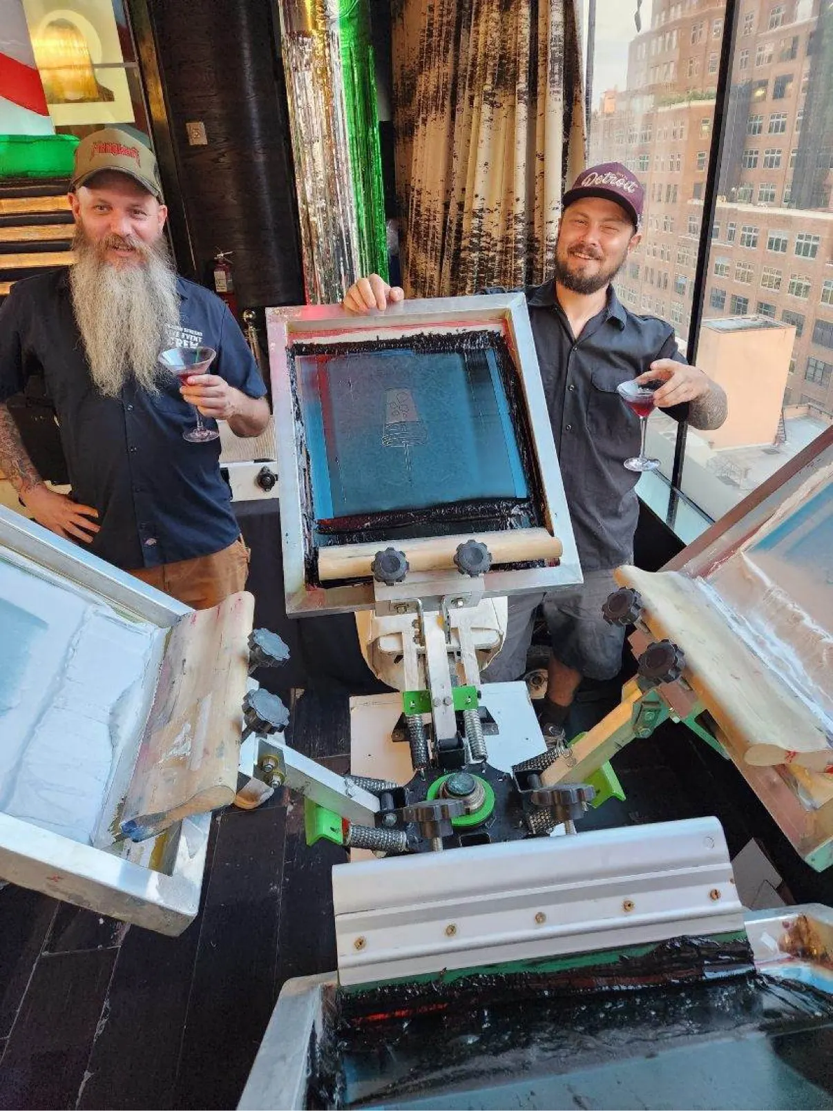 Two crew members posing with the press at a high-rise venue event, martini glasses in hand, city towers outside the windows