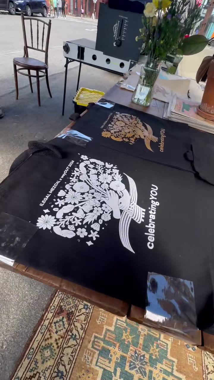 Black totes with a white botanical print laid out on the table at an outdoor storefront event, the dryer behind