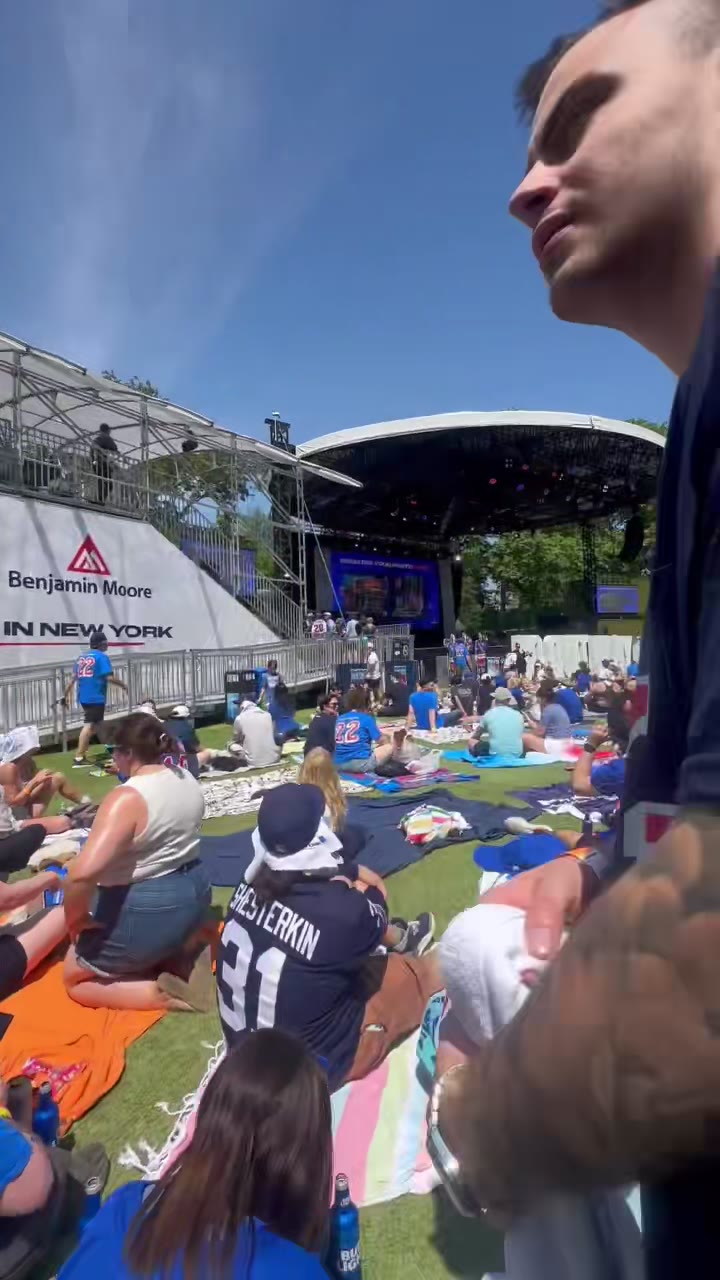 Fans in team jerseys spread across the lawn at an outdoor fan fest, watching the stage