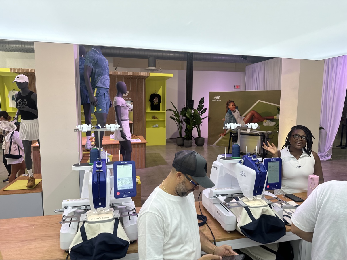 Two Brother embroidery machines stitching tote bags at the counter inside the New Balance Coco's Court tennis pop-up, the Coco Gauff poster wall behind