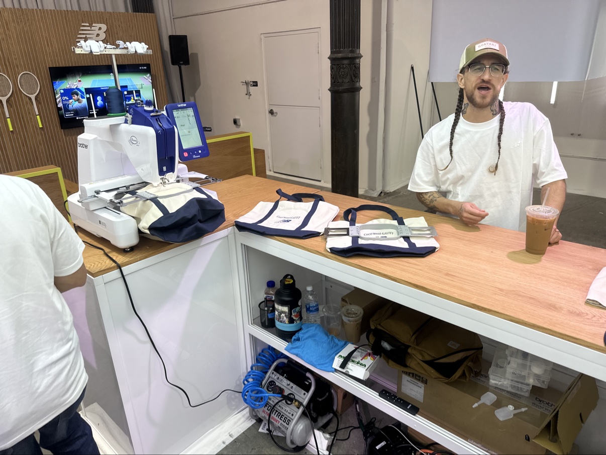 The counter at the New Balance Coco's Court pop-up — an embroidery machine running and finished embroidered totes lined up, a crew member behind the counter