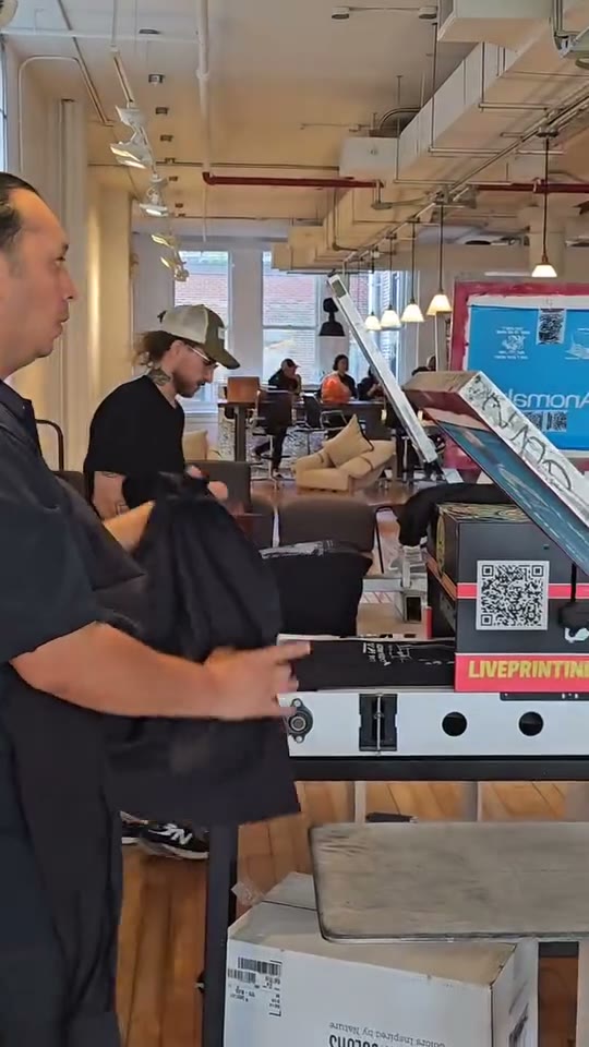 Their dryer with the Live Printing sticker and QR code at a loft event, a crew member holding a fresh black piece off the belt