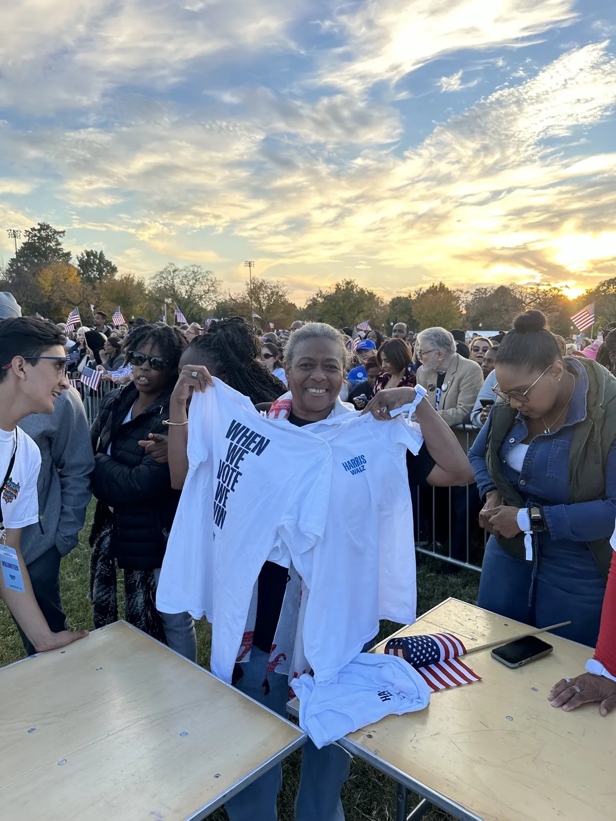 A guest at the DC rally holding up two fresh white tees at sunset, the crowd and flags behind her