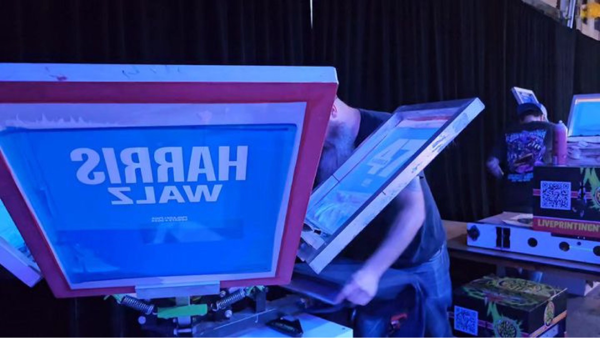 The Harris/Walz screen up close on the press, a crew member working behind it and their dryer with the Live Printing sticker at the side