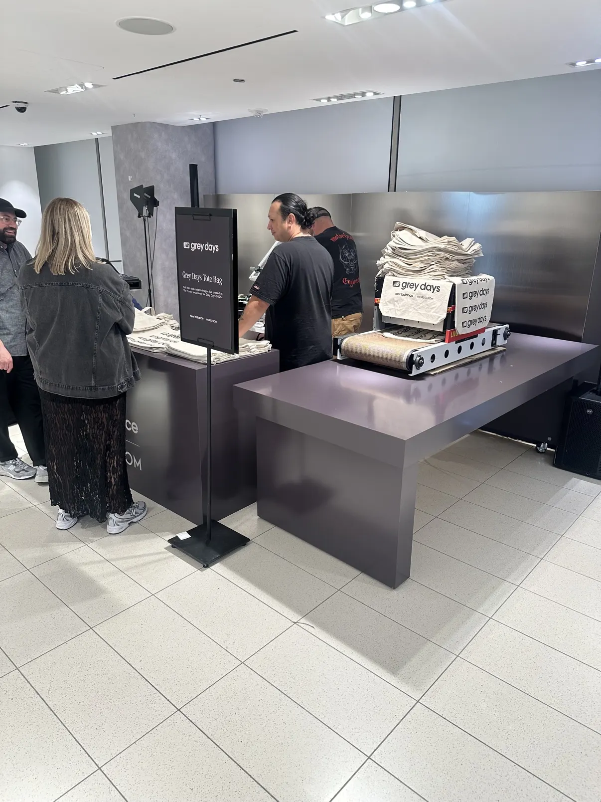The New Balance × Nordstrom Grey Days tote station inside Nordstrom — the branded counter and sign, their dryer stacked with Grey Days totes, guests at the counter