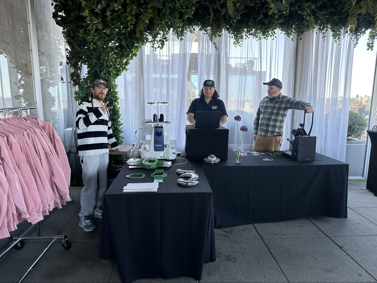 The embroidery and heat-press station on a greenery-draped rooftop terrace — Brother machine, laptop and letter samples on the tables, a rack of pink jackets ready to customize