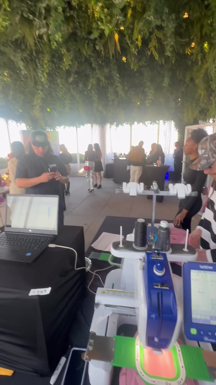 The rooftop embroidery machine mid-job, guests gathered around the tables under the greenery canopy