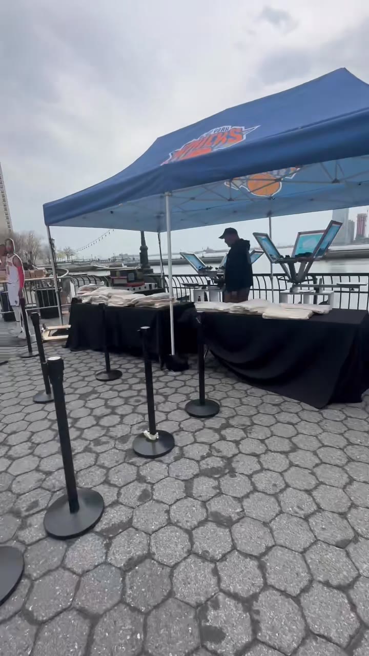 The Knicks tent station on the waterfront pier — press and tables set up, stanchion lanes laid out, the water behind
