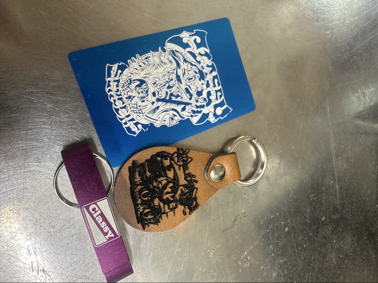 Finished laser-engraved pieces on brushed steel — a blue anodized card with their wizard art, a leather keychain with a hot-rod engraving, and a purple bottle-opener keychain engraved Classy