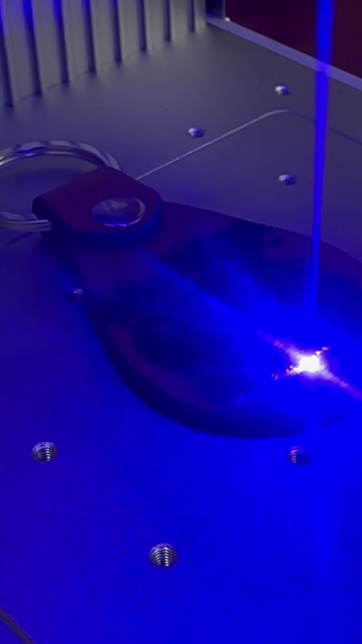 The open laser's beam sparking on a leather keychain mid-engrave, lit deep blue