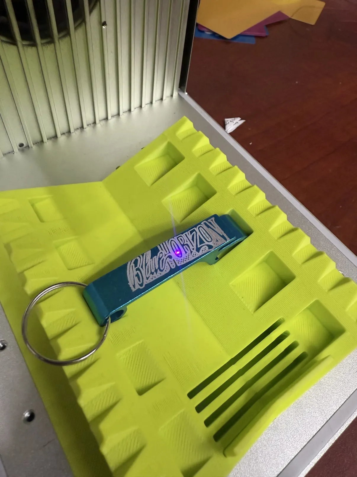 A teal bottle-opener keychain mid-engrave in the laser's neon jig, script lettering etching in with the beam dot on it