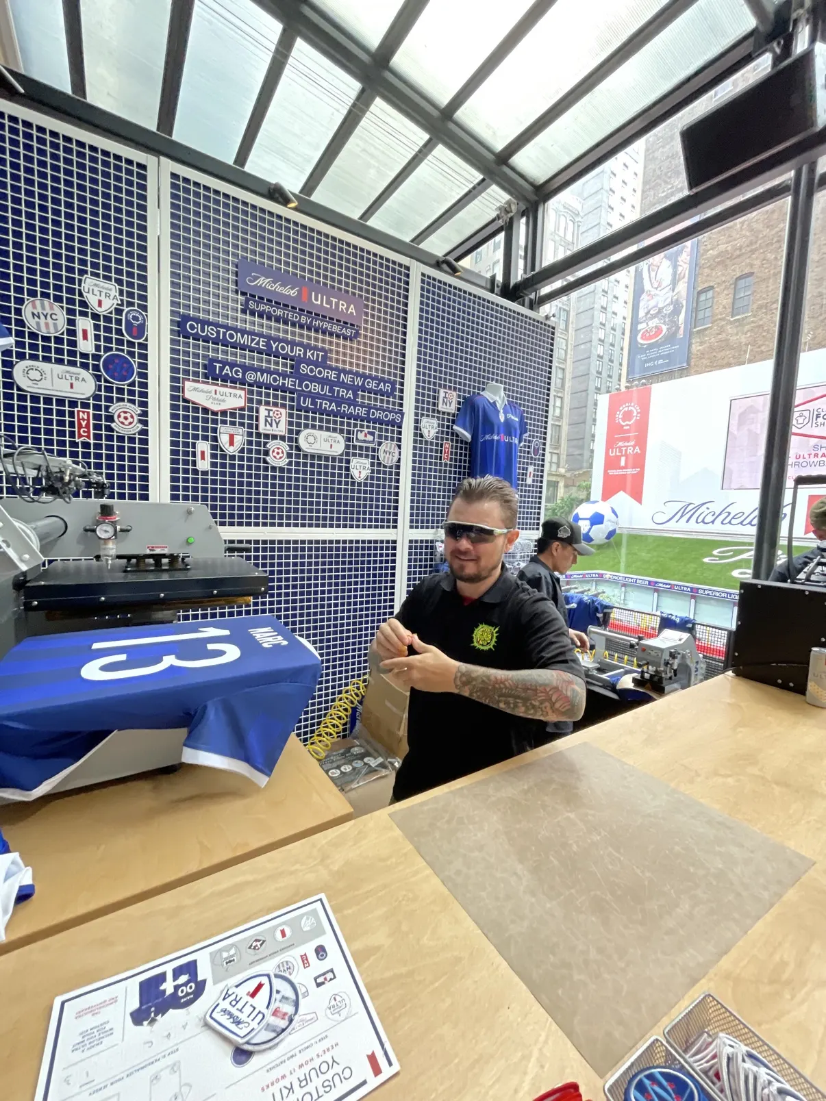 The Michelob Ultra × Hypebeast Customize Your Kit booth in full — the patch wall, a numbered soccer jersey on the press, the patch menu on the counter and the outdoor pitch visible through the glass