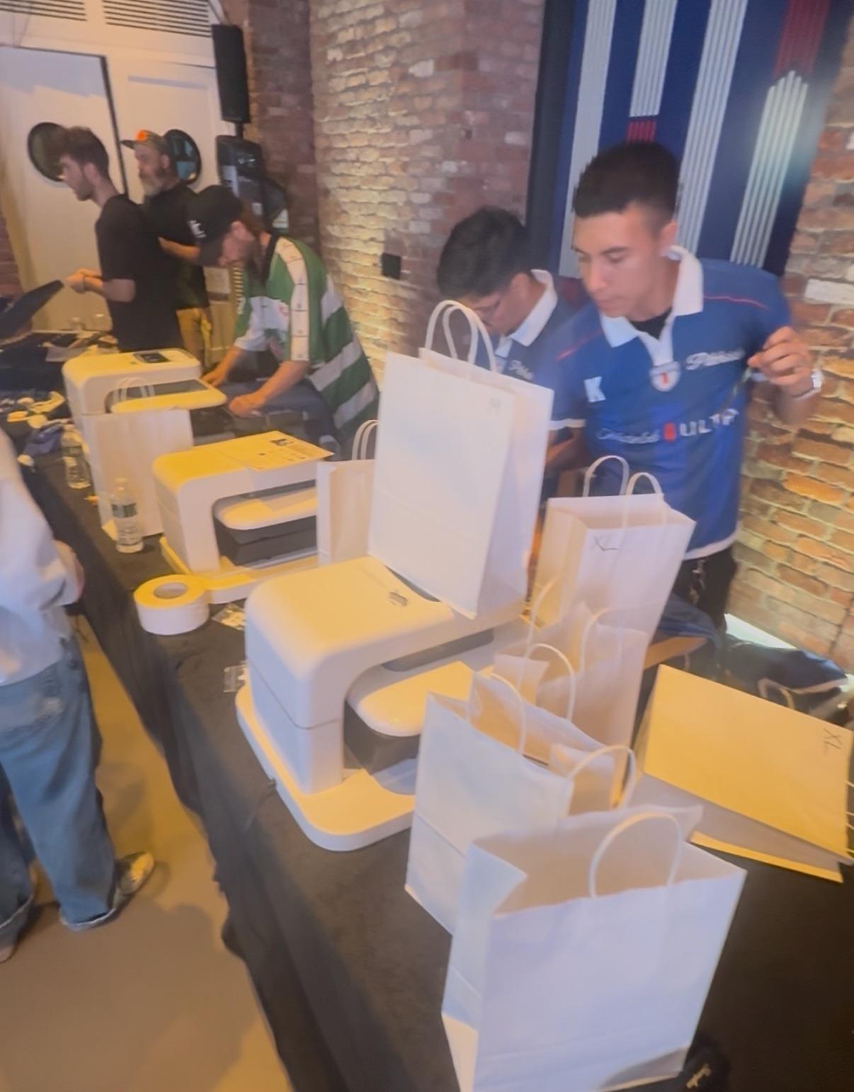 The three-laser event line from another angle — machines on the table and guests in team jerseys waiting with their bags